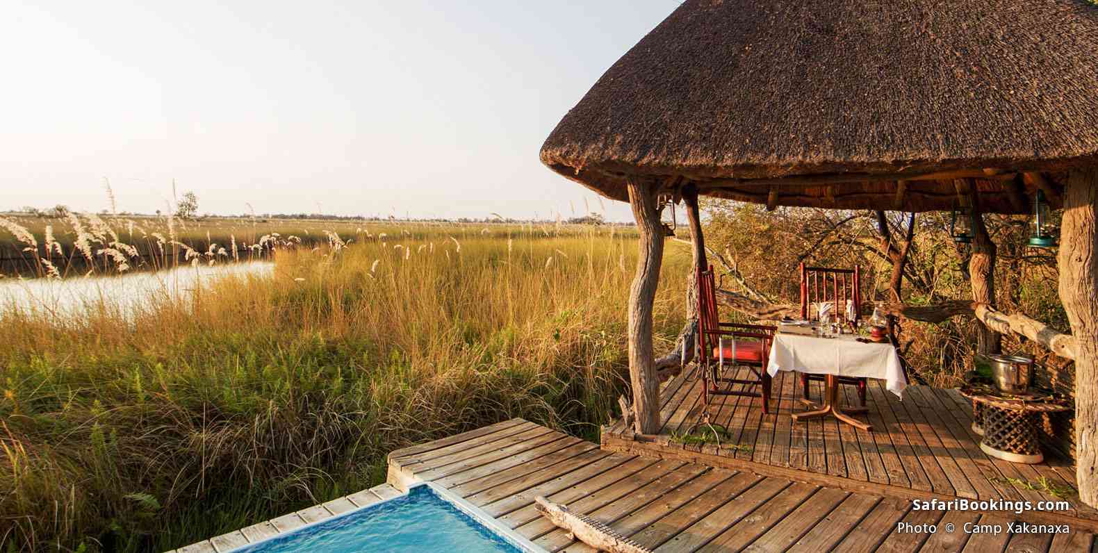 A rustic thatched-roof deck with a dining table overlooks tall grasses and a serene river beside a small plunge pool, Camp Xakanaxa